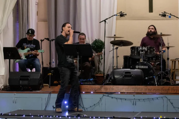 Band performing on stage with guitarist, vocalist, bassist, and drummer playing in front of draped curtains and lighting.