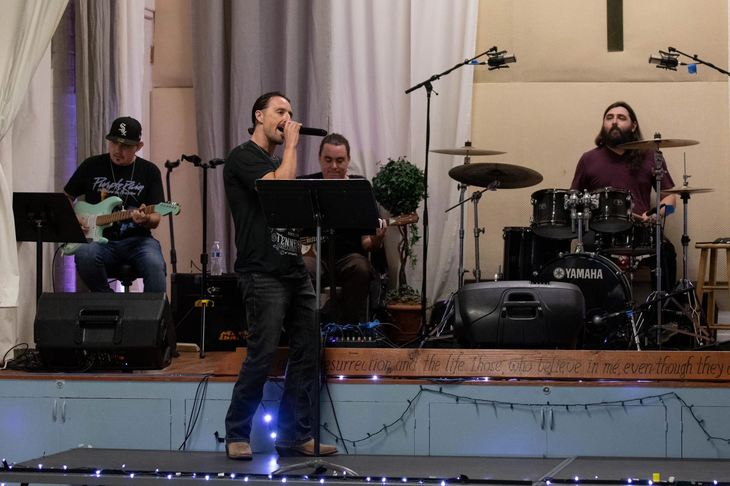 Band performing on stage with guitarist, vocalist, bassist, and drummer playing in front of draped curtains and lighting.